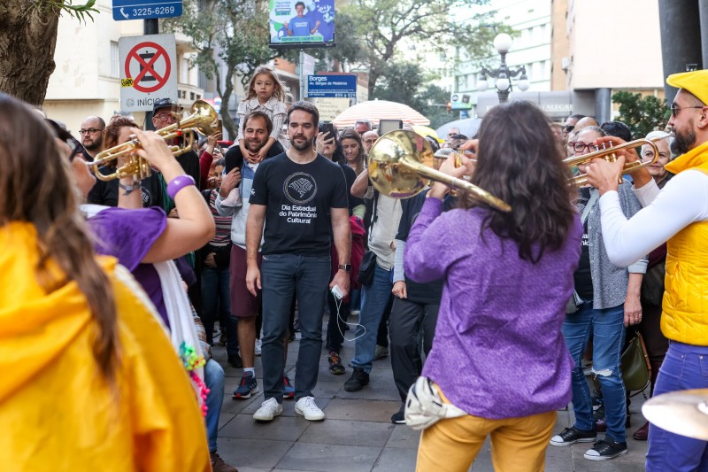 Governador Eduardo Leite, vestindo camiseta preta com a logo do Dia do Patrimônio, observa músico da fanfarra Bate & Sopra que tocam instrumentos de sopro diante do público do cortejo cultural que percorreu ruas do centro de Porto Alegre.
