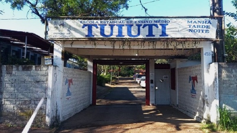 Imagem da entrada Escola Tuiuti em Gravataí. Do lado esquerdo podem ser vistas salas de aula e ao centro o portão de entrada na escola aberto. è uma área coberta e no alto há um painel com o nome da escola.