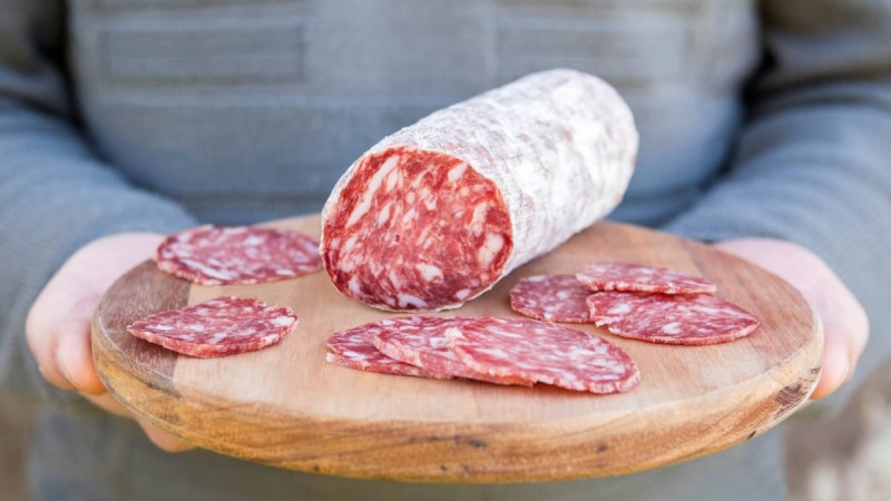 Uma pessoa segura um suporte de madeira em close que contém uma peça de salame com algumas fatias ao lado.