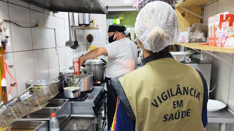 Numa cozinha, uma mulher aparece de costas. Em seu colete est&aacute; escrito Vigil&acirc;ncia em sa&uacute;de. Ela usa uma touca higi&ecirc;nica na cabe&ccedil;a. Na cozinha, h&aacute; um homem cozinhando junto ao fog&atilde;o. H&aacute; v&aacute;rias panelas, uma pia, e uma prateleira com alimentos no ambiente, que &eacute; apertado.