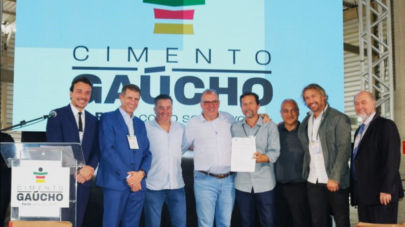 Foto posada em que aparecem o secretário Ernani Polo e o ex-jogador Lugano. Em um painel atrás dos oito homens que estão reunidos para a foto e no púlpito mais a esquerda da imagem pode ser vista a logomarca do cimento que será produzido com o nome Cimento Gaúcho.