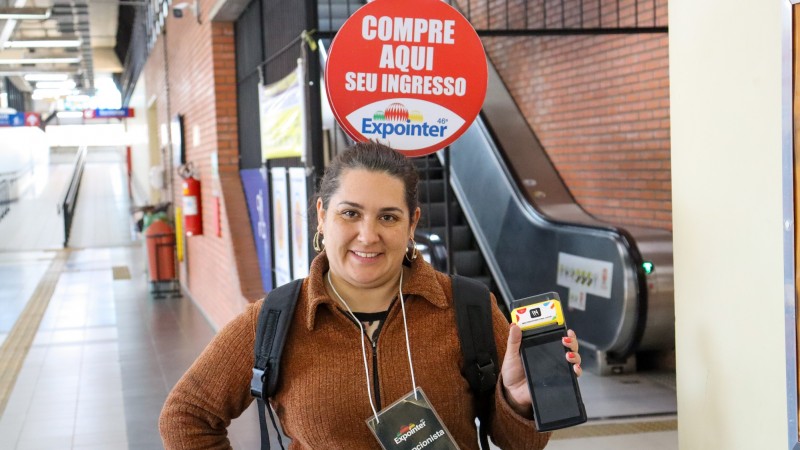 Numa estação de trem, uma mulher está de pé, em frente a uma escada rolante. Ela sorri. Está com uma mão na cintura e outra segurando uma máquina de cartão de crédito, que é exibida para a câmera. Ela está identificada com um crachá no qual se lê Expointer. Em suas costas, ela carrega uma mochila e uma placa que termina numa altura acima de sua cabeça, de cor vermelha; nela, lê-se Compre aqui seu ingresso, com a marca da Expointer logo abaixo.