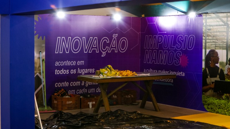 A imagem mostra um estande em processo de montagem. Há dois grandes painéis de cor azul escura, onde se lê INOVAÇÃO de um lado e IMPULSIONAMOS no outro. Há um mesa com alguns itens no meio. O chão está coberto por plástico preto. Não há pessoas na foto.