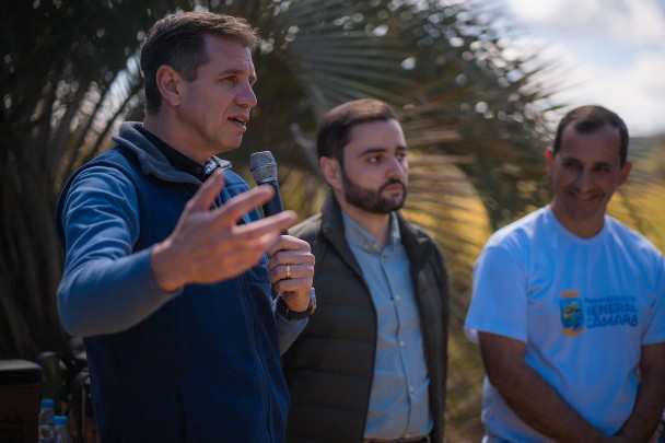 Secretário de Desenvolvimento Econômico, Ernani Polo, fala segurando um microfone com a mão direita enquanto gesticula com a mão esquerda. Ele veste camisa azul sob um colete azul marinho. Ao lado, o vice-governador Gabriel Souza.