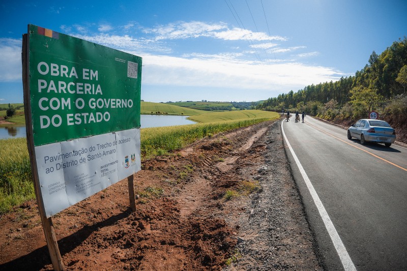 Placa com o texto "obra em parceria com o governo do Estado" aparece às margens da estrada asfaltada, no trecho que faz a ligação entre a ERS-244 e a vila de Santo Amaro do Sul, em General Câmara. Na pista da estrada, um carro branco. Ao fundo, árvores e o céu azul.