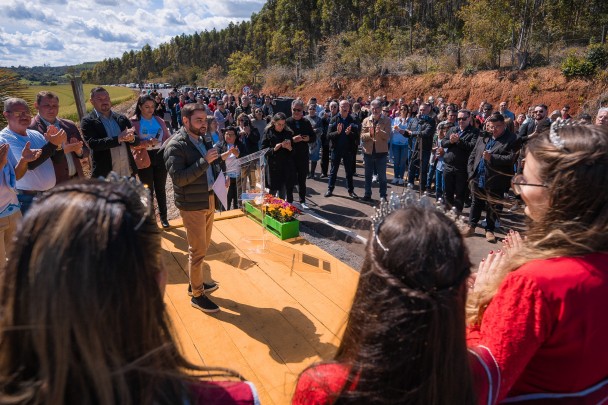 O vice-governador Gabriel Souza fala segurando um microfone sobre um palco de madeira às margens da via asfaltada que faz a ligação entre a ERS-244 e a vila de Santo Amaro do Sul, em General Câmara. Ao redor do palco, um grande grupo de pessoas da comunidade local observa.