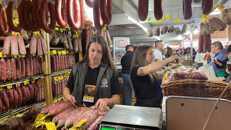 As irmãs Bini, de 20 e 23 anos, estão no comando de seus empreendimentos nos segmentos de embutidos e produtos lácteos. A foto mostra as duas gurias, uma com coleta cinza da emater e a outra de camiseta preta, ambas cabelo loiro castanho comprido, atrás de um balcão onde estão penduradas várias peças de salame.