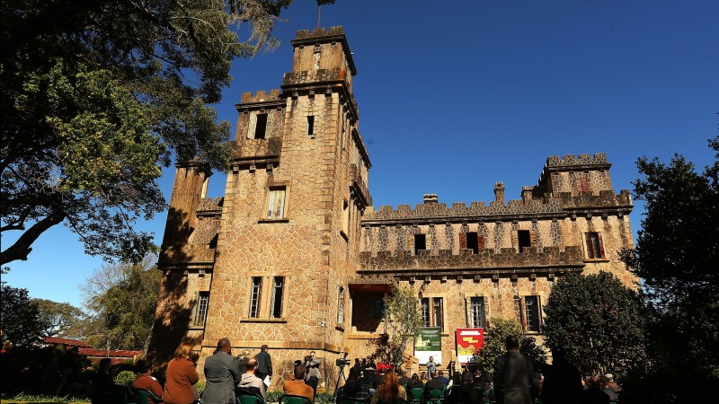 O Castelo de Pedras Altas aparece ao fundo. O dia está ensolarado e podem ser vistas pessoas, de costas, em pé e em cadeiras assistindo a alguém que fala na escadaria do castelo ao longe.