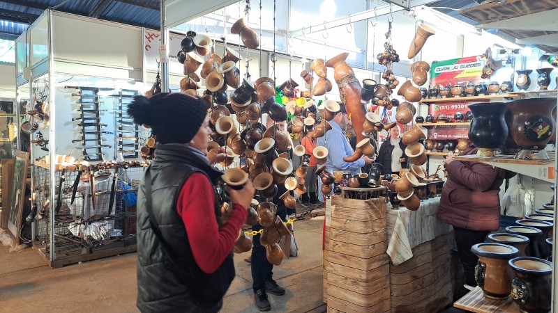Num galpão da feira, uma mulher de toca e colete para frio segura uma cuia. Ela está de lado para a câmera. À frente, está um estande que vende cuias, dispostas em prateleiras e penduradas. 