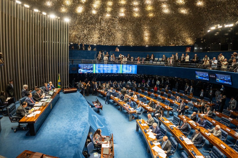 Fotografia lateral do senado. À esquerda, está a tribuna; à direta, o local onde ficam dispostas as longas mesas em que se sentam os senadores, no andar de cima, fica o espaço para quem quer acompanhar as sessões. O piso é azul.