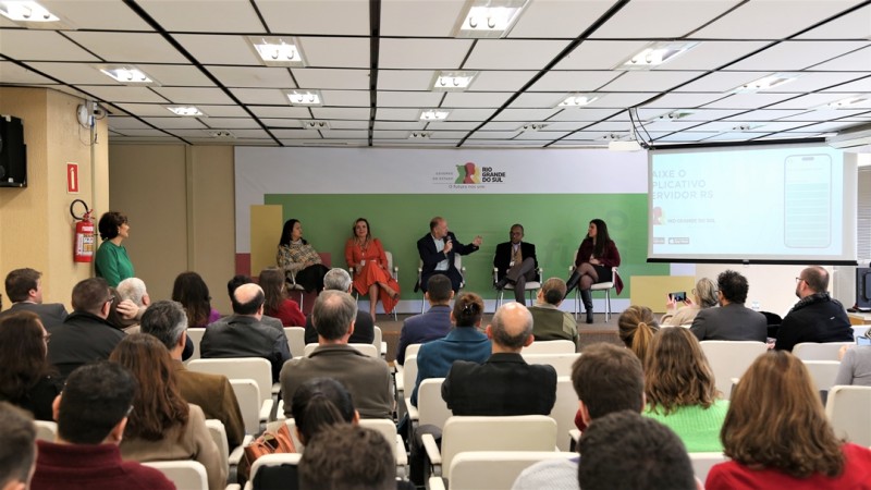 A imagem enquadra, a partir do fundo auditório, a frente do palco. Nele, estão sentadas cinco pessoas. A que estão no meio fala ao microfone e olha para o lado, apontando o slide. A plateia aparece na metade inferior da imagem, no primeiro plano, sentada em cadeiras de cor branca.