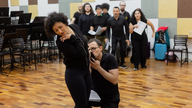 Numa sala de teatro, com piso de taco, há diversos atores ensaiando. Eles usam roupas pretas. No primeiro plano, há uma mulher de pé com rosto virado de lado e expressão sofrida; ajoelhado, está um homem que beija intensamente seu braço. Ao fundo, vários atores observam a cena, com expressão de espanto.