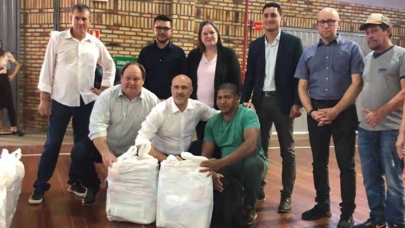 Numa quadra esportiva de piso marrom, várias pessoas posam para a câmera. A maior parte está de pé. Há três agachadas junto a sacolas com os alimentos doados.