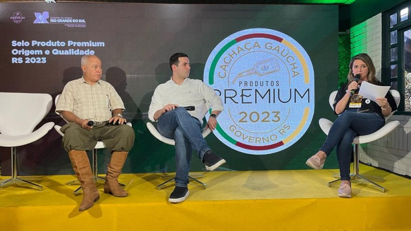 No palco do RS Innovation Agro, aparecem dois homens brancos sentados em cadeiras brancas. Um deles é um senhor grisalho, de camisa blaca, bombacha e bota. Outro é um homem mais hovem, de camisa branca, calça jeans e tênis. Ao lado deles, o selo lançado é mostrado em um telão. À direta da foto, uma mulher, também sentada, de camiseta preta, calça jeans e tênis. Ela fala ao microfone e segura um papel.