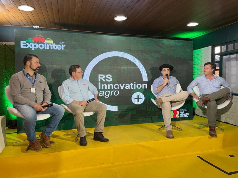 No palco do RS Innovation Agro, quatro homens participantes de um painel aparecem sentados em cadeiras brancas. Atrás deles, um telão com a logo do RS Innovation Agro.
