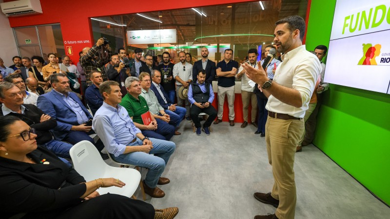 Numa sala fechada, Leite está de pé, à direita. Ele discursa para uma plateia cheia, sentada em cadeiras brancas. Ele gesticula com as mãos.
