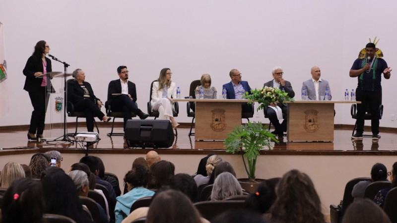 Num auditório, a foto enquadra, frontalmente, na parte inferior, a plateia e, na parte superior, o palco. No palco, há duas mesas e várias cadeiras, onde há pessoas sentadas participando do painel. Em cada ponta do palco há uma pessoa em pé. Á direita, um homem indígena fala ao microfone.