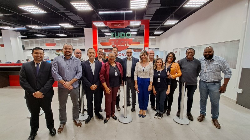 Na unidade do Tudo Fácil, que possui paredes e piso brancos, há um painel de vidro ao fundo com a logo verde e vermelha do Tudo Fácil. À frente, estão várias pessoas de pé posando para a câmera. Elas sorriem.