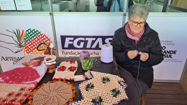 Num estande do pavilhão, há um painel baixo, de cor branca, com as logos do governo do Estado e da FGTAS. À frente, uma senhora idosa, bem agasalhada, está sentadas junto a uma mesa onde estão depositadas peças coloridas de crochê. Ela está fazendo chochê, e sorri.