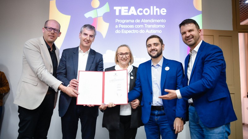 Numa sala fechada, os participantes do ato estão de pé e exibem o documento assinado, sorrindo para a foto. São quatro homens e uma mulher. Ao centro, está Arita. Ao fundo, está escrito Teacolhe, com a logo do programa.