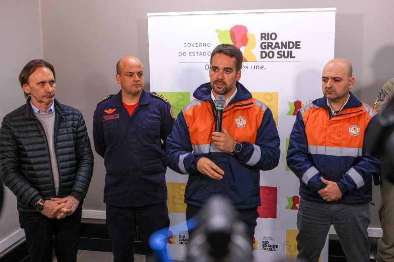 Num ambiente fechado, Leite e mais três homens estão de pé em frente a um painel com a logo do governo. O governador está com um blusão da Defesa Civil, de cor laranja. Ele fala ao microfone.