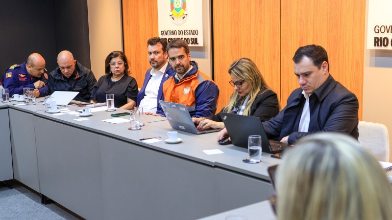 Em uma sala de reunião, Leite está sentado junto a uma mesa contínua com diversos secretários. Ele usa um colete laranja da Defesa Civil. Os secretários trajam roupas formais. Leite olha de lado para alguém que fala. Ao fundo, há um painel de cor marrom com placas onde está escrito "Governo do Estado do Rio Grande do Sul"