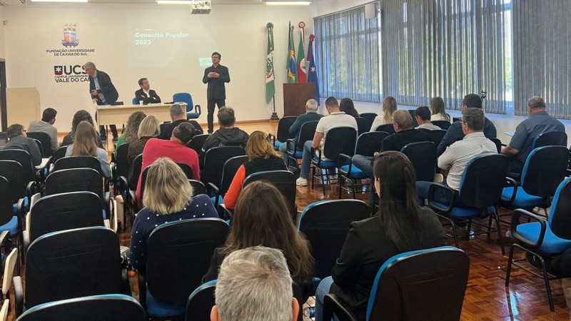 Num auditório de paredes brancas, piso de taco e janelas à direita (tapadas por persianas), várias pessoas estão sentadas em cadeiras azuis, assistindo a uma assembleia. O evento foi enquadrado do fundo, de modo que o público está em primeiro plano. Ao fundo, no palco, há três homens. Um está de pé e fala; outro esta se levantando de uma cadeira; e outro está sentado numa cadeira junto a uma mesa. À direita da fotografia, ainda no palco, há bandeiras do Brasil e do RS, além de mais duas (difíceis de identificar). Na parede ao fundo, está a logo da UCS escrita sobre os dizeres "Vale do Caí".