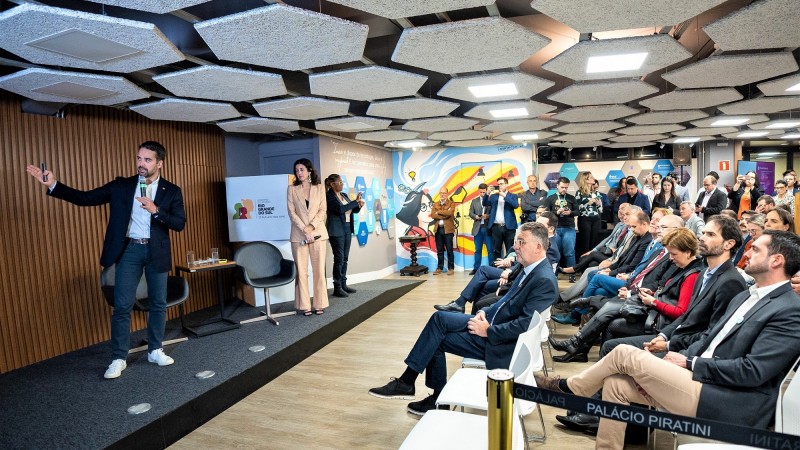 Leite em pé, mais à esquerda da imagem, falando à plateia com um microfone. À sua direita, em pé, Simone Stülp e à direita da imagem está a plateia assistindo à apresentação do governador.