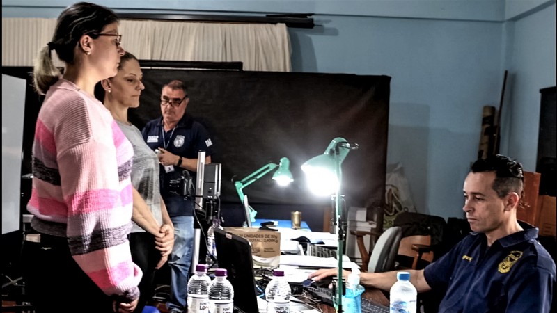 Duas mulheres do lado esquerdo da imagem diante de uma mesa, onde um servidor do IGP, do lado direito da imagem, sentado, trabalha ao computador. 