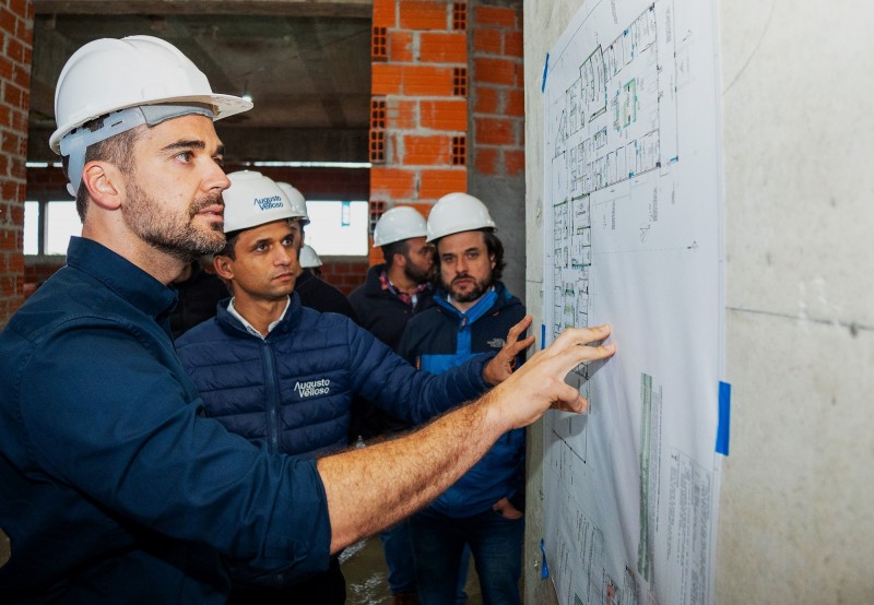 Leite de perfil no canto esquerdo da imagem com a mão sobre uma planta de engenharia afixada na parede à sua frente. Podem ser vistas outras pessoas ao fundo acompanhando as explicações dadas ao governador por um homem que está ao seu lado, de frente, também com as mão sobre a planta de engenharia.