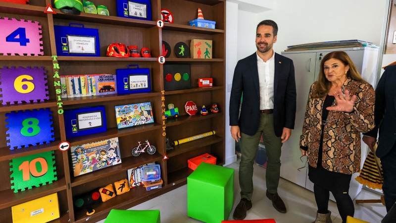 Leite em pé ao lado de uma mulher conhecendo uma sala da escola, na qual pode ser vista uma estante com diversos materiais utilizados em atividades educacionais.