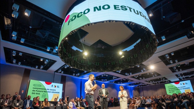 Leite no centro de um palco falando para plateia reunida em torno do palco. Acima, no teto, um grande painel circular onde se lê Foco no Estudante.