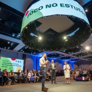 Leite no centro de um palco falando para plateia reunida em torno do palco. Acima, no teto, um grande painel circular onde se lê Foco no Estudante.