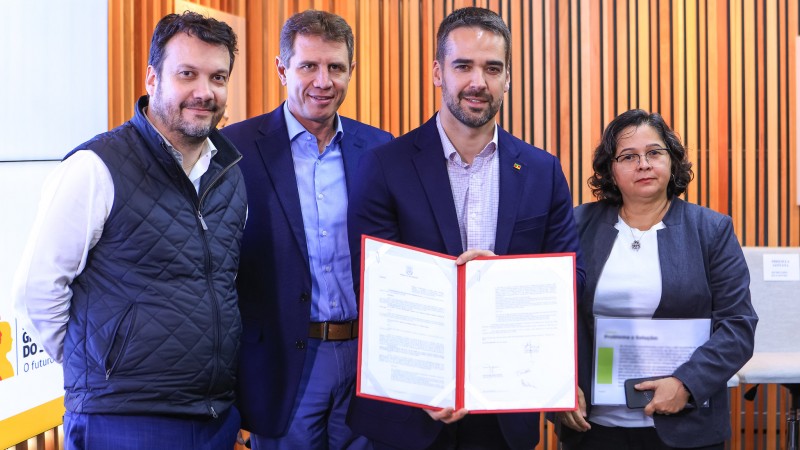 Numa sala cujas paredes são de cor madeira, as quatro pessoas posam para a foto, sorrindo e exibindo o papel assinado.