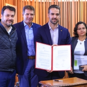 Numa sala cujas paredes são de cor madeira, as quatro pessoas posam para a foto, sorrindo e exibindo o papel assinado.