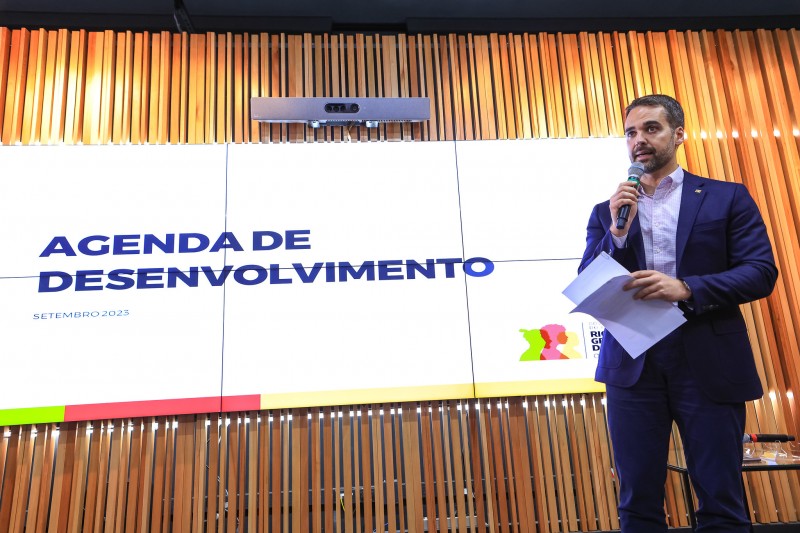 Em frente a um painel no qual está escrito Agenda de desenvolvimento, Leite está de pé e fala ao microfone, segurando também alguns papeis. Ele está à direita na foto.