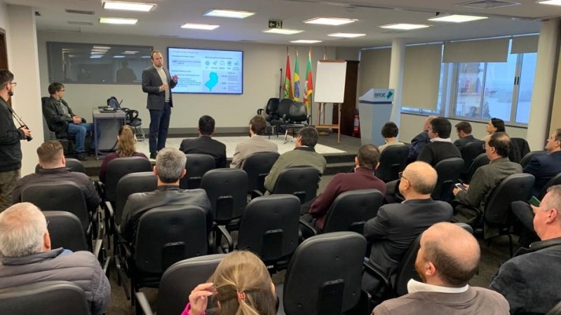 Foto de um evento em que se vê pessoas de costas em uma plateia e ao fundo um homem de pé fazendo uma apresentação sobre um tablado e com uma tela atrás de si.