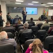 Foto de um evento em que se vê pessoas de costas em uma plateia e ao fundo um homem de pé fazendo uma apresentação sobre um tablado e com uma tela atrás de si.