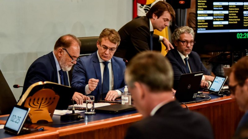 Imagem aproximada da Mesa da Assembleia durante a votação do PPA 2024-2027. Podem ser vistos dois deputados sentados conversando, um em pé e outro sentado olhando para frente.
