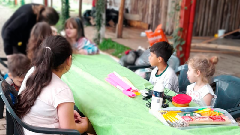 Em ambiente externo, seis crianças estão sentadas ao redor de uma mesa. Uma pessoa adulta conversa com uma das crianças, na ponta da mesa. Há materiais escolares para atividades em cima da mesa. 