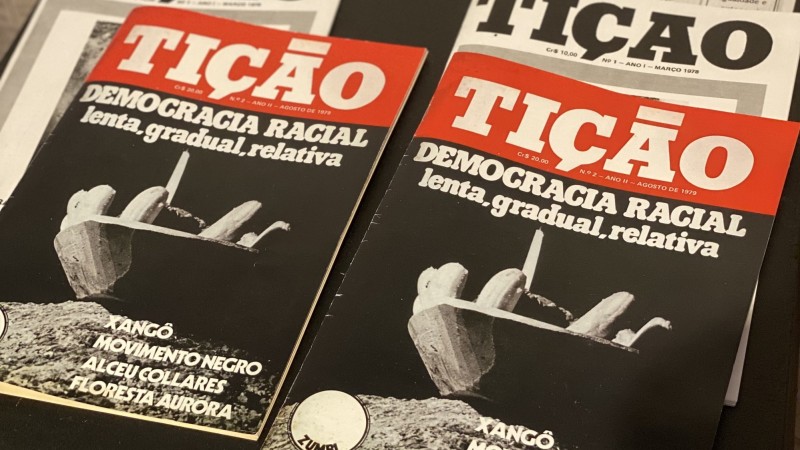 Dois exemplares da revista estão dispostos numa mesa. Tição está escrito no topo, em letras brancas contra fundo vermelho. A fotografia de capa é preto e branca, e o conteúdo é difícil de identificar. Está escrito: "Democracia racial - lenta, gradual, relativa".