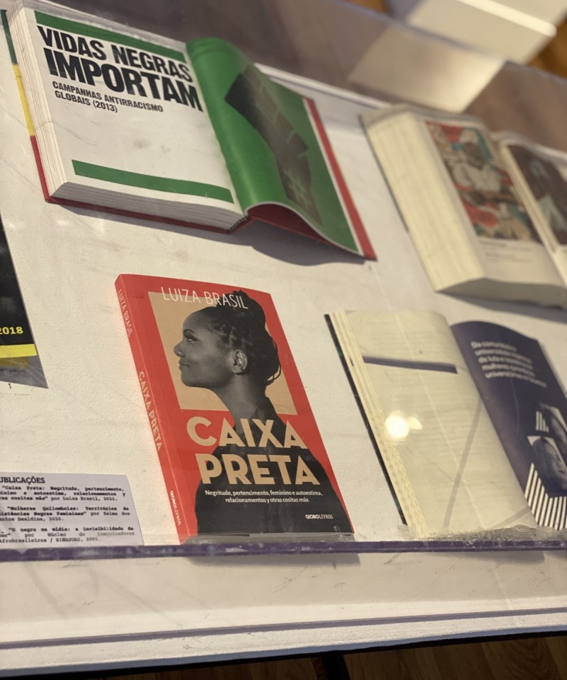 Numa mesa coberta com vidro, publicações sobre a temática negra. Um livro aberto apresenta os dizeres "Vidas negras importam"; outro, de capa vermelha, tem como título "Caixa Preta".
