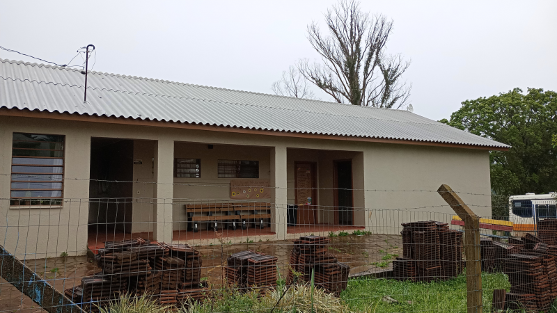 Fachada de uma escola cujas cujas paredes e telhas são claras. Há uma cerca simples de arame no primeiro plano. No pátio da escola, na grama, há materiais de construção empilhados. O céu está nublado.