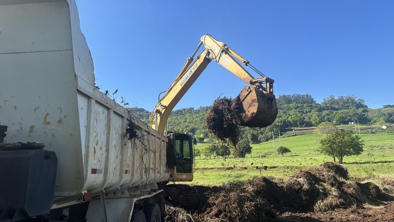 Imagem de uma caminhão com uma retroescavadeira jogando terra na sua caçamba no início da terraplanagem da área para construção de casas provisórias em Arroio do Meio.