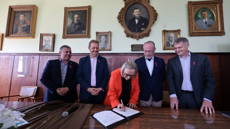 Numa sala em que há pinturas penduradas nas paredes, quatro homens em trajes formais aparecem de pé, aguardando enquanto a secretária Arita assina um documento, apoiado numa mesa de madeira. Ela está ao centro, entre os demais participantes da solenidade.