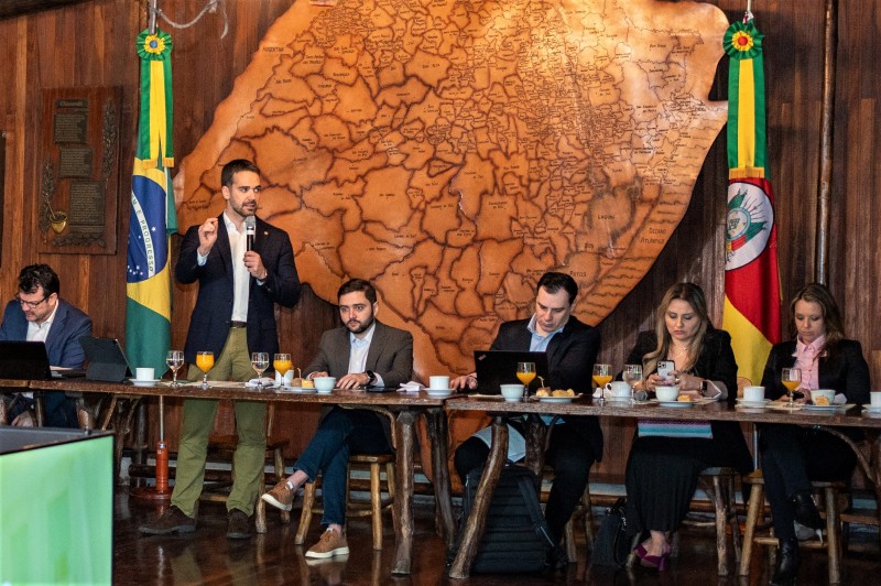 Leite em pé com o microfone falando durante uma reunião. Ele está atrás de uma mesa e à qual estão sentados um homem à sua esquerda e dois homens e duas mulheres à sua direita. Ao fundo, da esquerda para direita: uma bandeira do Brasil, o mapa em madeira do Rio Grande do Sul e uma bandeira do Rio Grande do Sul.