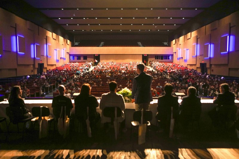 Foto em ambiente fechado, mostra os integrantes da mesa de abertura do evento de costas. Ao fundo, está o público da conferência, sentado em cadeiras do auditório.