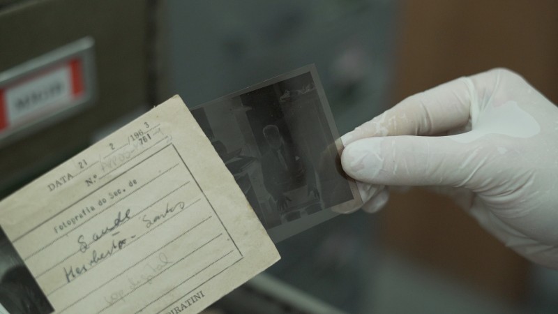 A foto enquadra uma mão, com luva cirúrgica branca, segurando uma ficha de papel antiga e um negativo de fotografia.