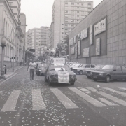 Foto em preto-e-branco mostra a rua em frente ao palácio. Há uma fileira de carros da época (aparentemente nos anos 1980) logo antes da faixa de pedestres. O primeiro da fila está com coberto com faixas. Uma pessoa caminha acompanhando os carros. 