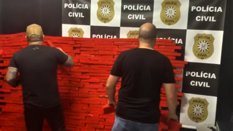 A imagem traz dois homens de costas diante de uma pilha grande de drogas apreendidas, embaladas como tabletes vermelhos. Ao fundo um painel quadriculado que traz distintivos da polícia civil na parte branca e e as palavras polícia Civil na parte preta.